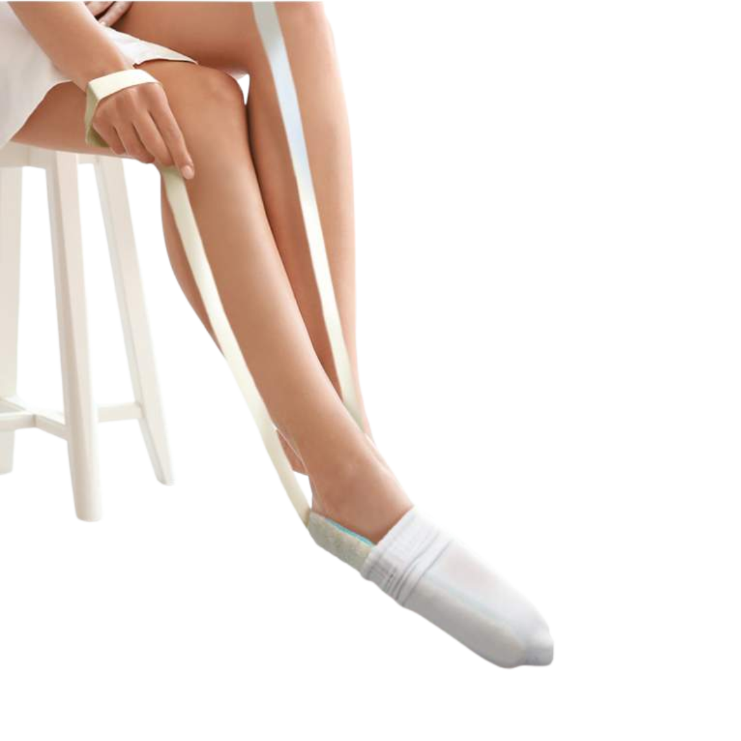 A person sitting on a stool demonstrating the use of the sock aid, pulling the white straps to slide a white sock onto their foot.