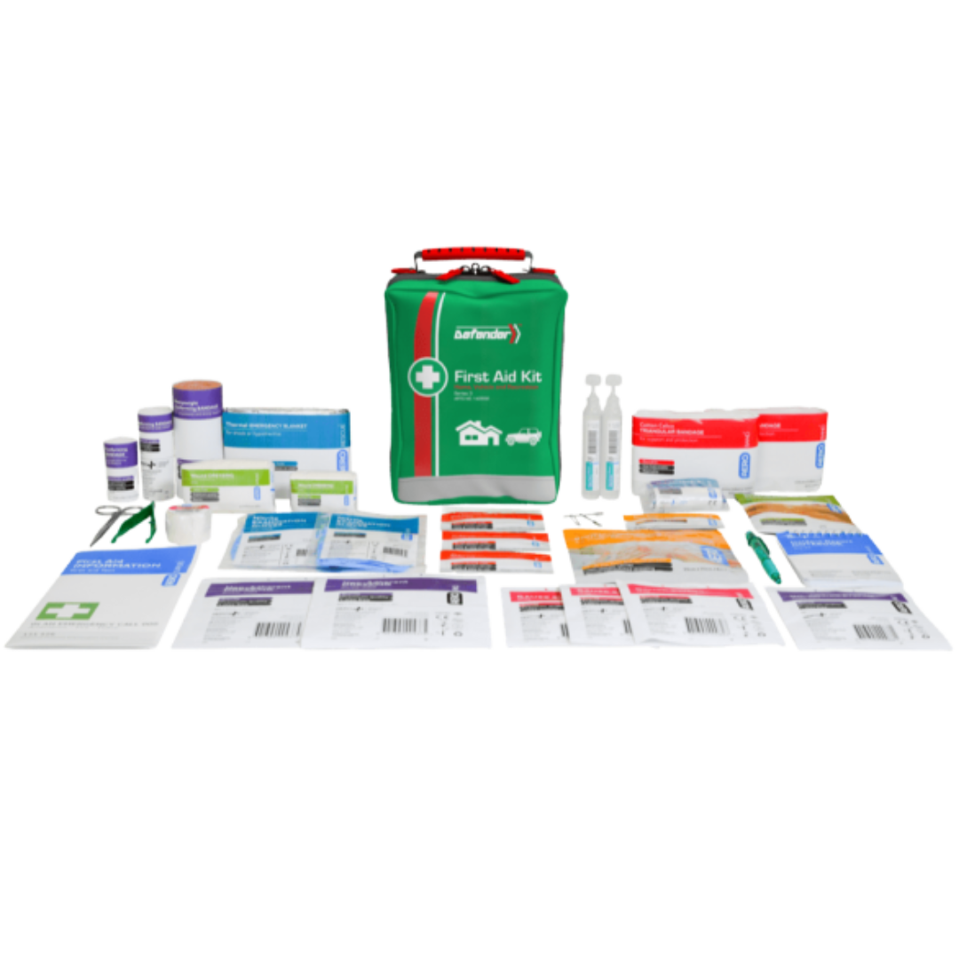 A product image showing the contents of the Defender 3 Series Softpack First Aid Kit laid out on a dark background. The green softpack bag is centered, surrounded by items including rolls of bandages, small liquid bottles or ampoules, a pair of scissors, a booklet, and various packaged wound dressings.